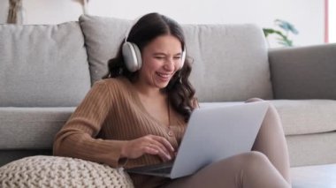 Kadın yerde oturuyor, parmakları dizüstü bilgisayarın klavyesinde dans ediyor ve içten bir kahkahayı paylaşıyor. Gözleri neşeyle parıldıyor ve kahkahalar odayı sıcaklıkla dolduruyor..