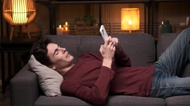 若い大人の男はソファーの上に横たわり ヨウを放り出しながら電話で魅了される シーンはリラックスした夜をキャプチャし テクノロジーの使用をブレンドし 居間の快適さのヒント — ストック動画