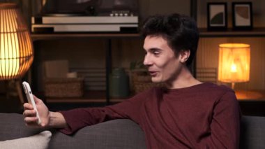 Genç, beyaz bir adam kanepede otururken telefonla görüntülü konuşma yapıyor. Oturma odası bu sanal bağlantı için rahat bir zemin sağlıyor, bir rahatlama anı yakalıyor..