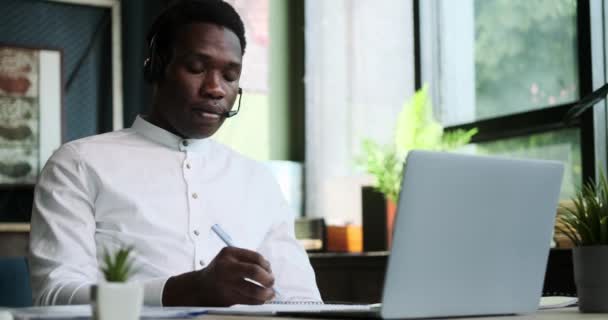 アフリカ系アメリカ人男性 ビジネスマン またはヘッドセット携帯電話を持つ学生 ラップトップでオフィスで勉強しています オンラインカスタマーサポートワーカー 学生リモートスタディ ビデオ通話への参加 — ストック動画