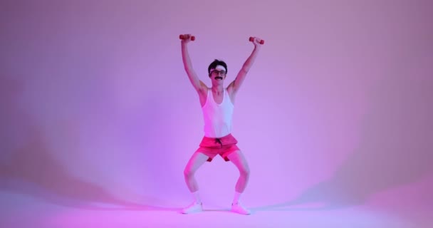怪异的 复古的80多岁的白人男子穿着运动服 站在紫色的背景上 用哑铃做蹲式运动 — 图库视频影像