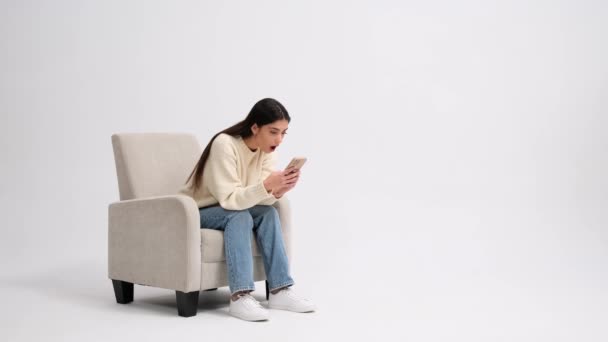 拥有手机的高加索女人对坐在扶手椅上的白色背景的新闻 销售或网上购物感到惊讶 社交网络 远程通信 在线技术 — 图库视频影像
