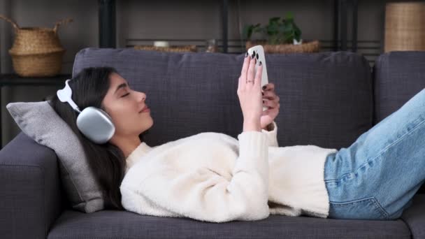 年轻的白人妇女拿着电话在客厅的沙发上休息 听着音乐 享受闲暇时间 在社交网络上度过周末或假期 — 图库视频影像