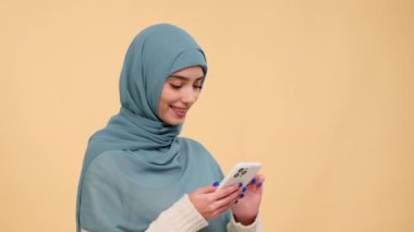 Tesettürlü pozitif Müslüman kadın internette sörf yapmak, internetten alışveriş yapmak, sosyal ağlarla iletişim kurmak, gülmek, olumlu tepki vermek, bej bir arka planda dinlenmek için telefonunu kullanıyor..