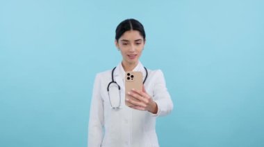 Pozitif bir kadın doktor ya da hemşire portresi mavi arka planda telefonda video konferansı yoluyla konuşuyor. Uzaktan hasta konsültasyonu, ilaç ve teknolojinin karışımı, modern sağlık hizmetleri.