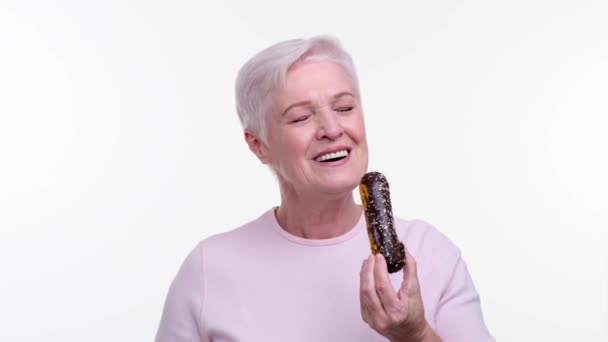 Sorridente Donna Anziana Caucasica Morde Felicemente Eclair Cioccolato Uno Sfondo — Video Stock
