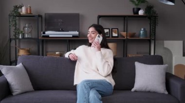 Tatmin olmuş, kaygısız, beyaz bir kadın akıllı telefonla konuşuyor ve oturma odasındaki kanepede otururken gülüyor. Çevrimiçi iletişim, evde dinlenme.