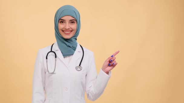 Portret Uśmiechniętej Muzułmańskiej Lekarki Wskazującej Palcem Prawo Beżowym Tle Pielęgniarka — Wideo stockowe