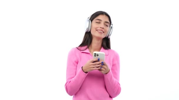 年轻的白人女人 戴着耳机 听音乐 用手机上网或上网娱乐 在白人背景下跳舞 — 图库视频影像