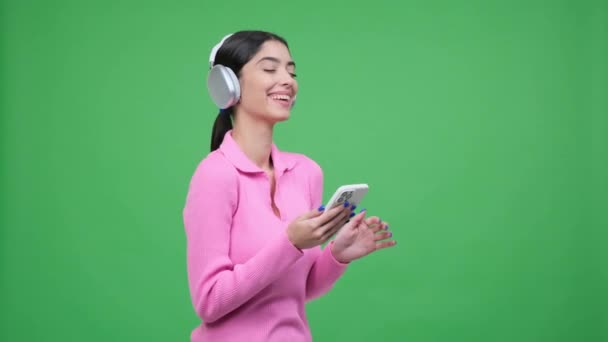 积极的白人年轻女性在社交媒体上用电话留言 用耳机听音乐 站在绿色背景上 — 图库视频影像