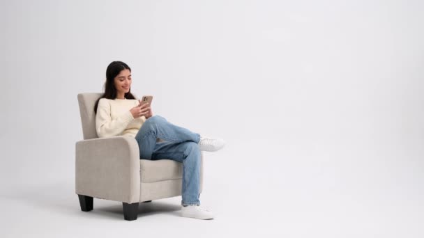 インターネットブラウジング オンラインショッピング エンターテイメントに携帯電話を使用して 白い背景にアームチェアに座っている友好的な白人女性 コピースペース — ストック動画