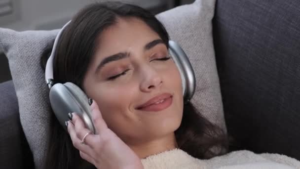 坐在客厅沙发上听音乐的戴着耳机的年轻而轻松的白人女性的近照 梦中的女人 音乐的幸福 — 图库视频影像