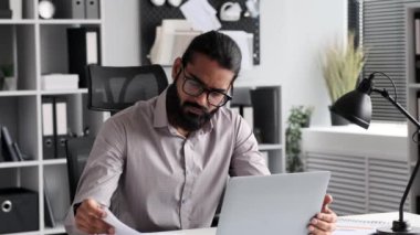 Sert Hintli erkek CEO belgeleri ve raporları kontrol eder, bir geliştirme stratejisi oluşturur, vergileri hesaplar, modern bir ofiste dizüstü bilgisayarla masa başında evrak işlerini halleder..