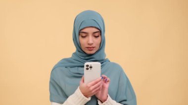 Bej arka planda cep telefonu kullanan konsantre olmuş Müslüman bir kadının portresi. İnternette gezinmek, sosyal medyada mesajlaşmak, telefonla dinlenmek..
