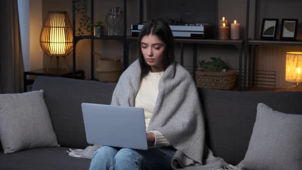 夜のリビングルームでソファーに座って リモートワーク ライティングストーリー ロマンス 出版物にノートパソコンを使用して 毛布に焦点を当てた若い女性 メールでの回答 — ストック動画