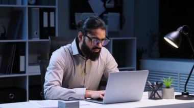 Ciddi Hintli erkek yönetici akşam ofisinde evrak ve dizüstü bilgisayarla çalışıyor. Proje geliştirme, finans ve bütçe planlaması, organizasyon vergileri.