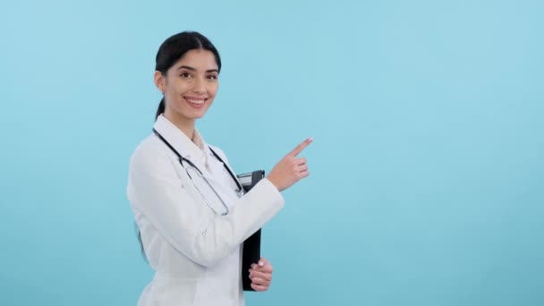 积极的医院工作人员或带着微笑的医生在蓝色背景的复制空间向右指了指 课本空间 年轻女医生 积极保健 — 图库视频影像