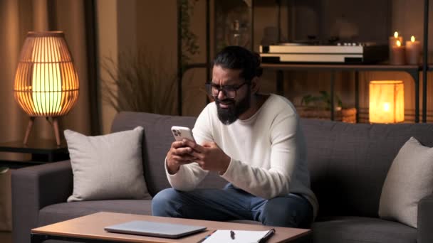インド人の男性は 夕方に家でソファーに座っている間 電話でインターネットを閲覧しながら休んでいます オンラインソーシャルメディア リラックスしたレジャー活動 — ストック動画