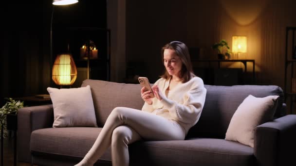 ポジティブな白人女性は 家庭のリビングルームで夕方にソファーでリラックスして電話を使用しています ソーシャルメディアアプリでのメッセージング インターネット閲覧 — ストック動画