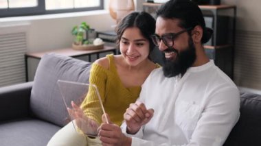 Pozitif Hintli çift, evdeki oturma odasındaki kanepede dinlenen online eğlence için şeffaf cam tablet bilgisayar kullanıyor. Modern dijital tablet şablonu.