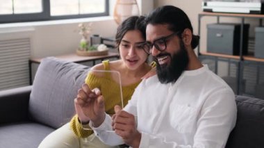 Mutlu Hintli çift koltukta modern dijital tabletle rahatlıyor. Çevrimiçi alışveriş, sosyal medya kullanımı, boş zaman aktivite konsepti. Gelecekçi aygıt şablonu.