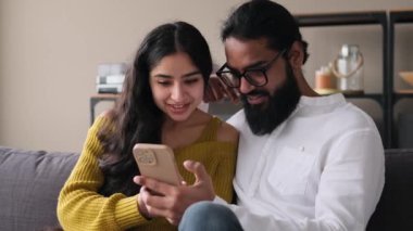 Genç mutlu Hintli çift boş zamanlarını telefon kullanarak evde oturma odasındaki kanepede dinlenerek geçiriyor. Çevrimiçi eğlence, bağlantı ve iletişim konsepti.