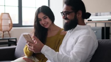 Mutlu Hintli karı-koca telefonla dinleniyor ve evdeki kanepede gülüyor. Eğlenmek, sosyal medyada sohbet etmek, birlikte vakit geçirmek..