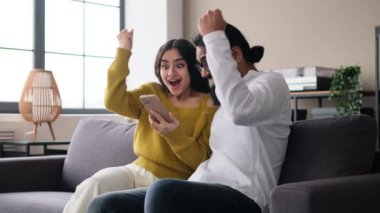 Aile şoke olmuş Hintli çift telefon kullanıyor, iyi haberler alıyor, başarıyı kutluyor, ev oturma odasındaki kanepede birbirlerine beşlik çakıyorlar..