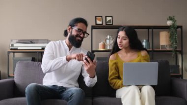Pozitif Hintli çift evlerinde cep telefonu ve dizüstü bilgisayar kullanarak cihazlarla kanepede dinleniyor ve gülüyorlar. Modern teknolojiyle rahatlıyor..