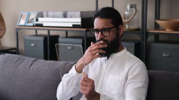 自宅でソファーで透明なタブレットコンピュータでオンラインで働く深刻なインドのビジネスマン リモートワーク 現代技術 デバイステンプレート — ストック動画