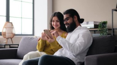 Şaşırmış Hintli karı koca telefon kullanıyor oturma odasında dinleniyor, iyi haberler alıyor ve haykırıyor, çevrimiçi başarıyı kutluyor..