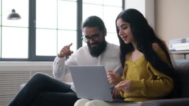 Pozitif Hintli çift evde kanepede otururken dizüstü bilgisayar kullanıyor, müzik dinliyor, birlikte şarkı söylüyor ve dans ediyor. Ev içi eğlence etkinliği konsepti.