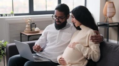 Hintli koca ve hamile eş oturma odasında kanepede otururken online alışveriş için dizüstü bilgisayar kullanıyorlar. Çocuk için hazırlık, doğum beklentisi..