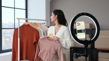 Arkadaş canlısı moda kadını Kafkas blogcu kıyafetleri satmak, kayıt yapmak veya sosyal medyada yayın yapmak için depoda duruyor. Moda görünümü oluşturuluyor.