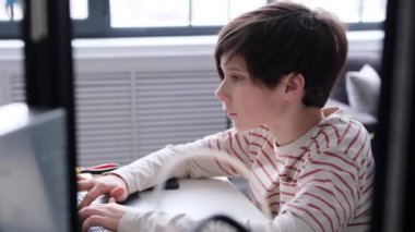 Ciddi bir beyaz çocuğun portresi e-öğrenim ve 3D yazıcı ve dizüstü bilgisayarla çalışıyor, atölyede masa başında oturuyor. Öğrenci bilgisayar kodu yazıyor..