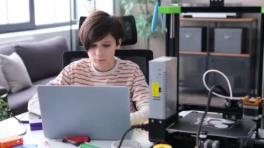 Yoğunlaşmış çocuk masada oturuyor, dizüstü bilgisayar ve 3D yazıcı ile ödev yapıyor. Proje araştırması, prototip icadı. E-öğrenme kavramı.