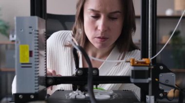 Odaklanmış beyaz kadın tasarımcı. 3D yazıcının çalışmalarını inceliyor ve izliyor. Cihazı kuruyor, yedek parça üretiyor. Ev atölyesi, DIY kavramı.