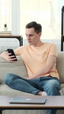 Genç beyaz adam koltuğa rahatça oturur ve mutluluk saçar. Yüzünde sıcacık bir gülümsemeyle, dijital dünyayla tamamen meşgul olan akıllı telefonuyla kolayca yön buluyor..