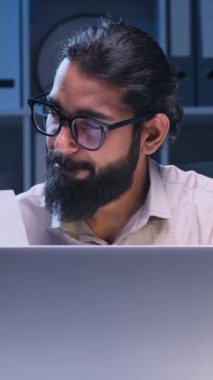 Hintli işadamı özenle kağıtlar ve bir dizüstü bilgisayarla çalışırken yakalandı. Profesyonel olarak giyinmiş, odaklanmış ifadeler geç saatlerde verimlilik ve adanmışlık hissi veriyor..