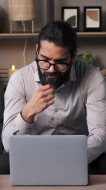 Oturma odasındaki kanepede oturan Hintli adam laptopta etkili bir şekilde çalışıyor. Odaklanmış ifadeler, iş ve ev hayatının kusursuz entegrasyonunu vurgulayarak, verimlilik ve uyum sağlayabilirliği iletir..