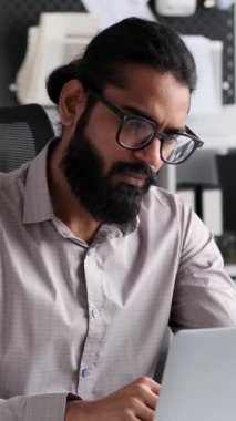 Ciddi bir Hintli işadamı, ofis ortamında dizüstü bilgisayara odaklanıyor. Uygulanan davranış ve kendini teknolojiye adamış olma, çalışmaya olan bağlılığını gösteriyor..