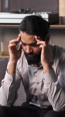 Hintli işadamı, oturma odasındaki bir kanepede oturuyor, baş ağrısıyla boğuşuyor, belki de iş baskısından. İfadeler rahatsızlık ve iç gözlem iletir.