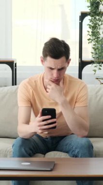 Evdeki kanepede oturan adamın yüzü keder ve hayal kırıklığıyla doluydu. Telefonu elinde, ekrana bakıyor, kendi düşüncelerinde kaybolmuş gibi görünüyor..