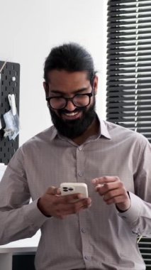 Gülümseyen Hintli işadamı, ofiste rastgele telefonuna göz atıyor. İfadeler, iş ve kişisel anlar arasında uyumlu bir denge ortamı yaratarak bir rahatlama ve rahatlama hissi verir..