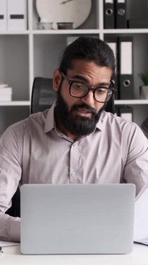 Hintli işadamı video görüşmesi için dizüstü bilgisayar kullanıyor ve evrak işleriyle uğraşıyor. Resmi bir kıyafetle süslenmiş, odaklanmış ifadeler modern teknoloji kullanımı ve geleneksel iş uygulamalarının bir karışımını iletir..