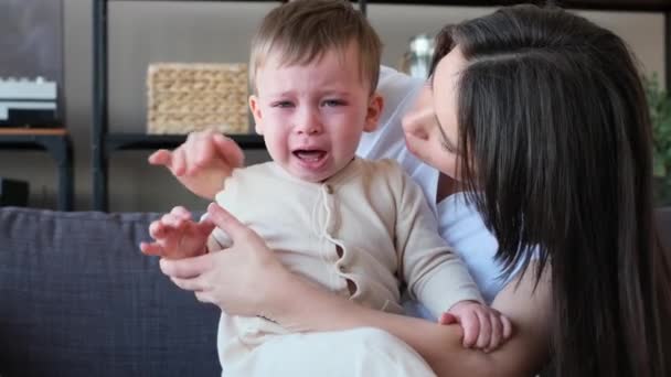 小男孩在家里客厅的沙发上一边哭一边用妈妈的手尖叫 妈妈抱着蹒跚学步的孩子 抚慰着他的头 抚摸着他的头 — 图库视频影像