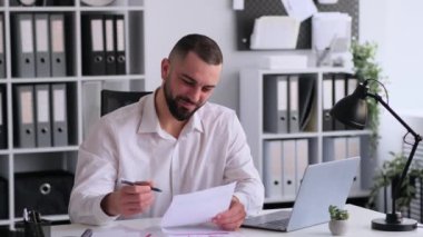 Dost canlısı Kafkas erkek ofis çalışanı kağıtlarla çalışıyor, bütçe planlıyor, analiz ediyor ve iş projelerini araştırıyor, ofiste masa başında oturuyor..