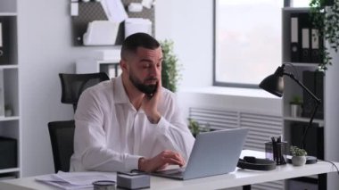 Çok çalışan Kafkasyalı iş adamı, masada dizüstü bilgisayarla çalışıyor, ofiste gözleri ovuşturuyor. Yorgunluk ve can sıkıntısı kavramı. Fazla mesai ve tükenmişlik.