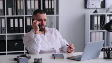 Gülümseyen Kafkas erkek ofis çalışanı teneffüste dinleniyor ve telefonla konuşuyor, modern ofiste masa başında oturuyor. Arkadaşlarla tatil planlamak.