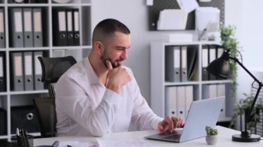 Laptop ve kağıtlarla çalışan, modern ofisteki masada oturan gülümseyen beyaz iş adamı. İş muhasebesi, bütçe analizi veya raporlar.